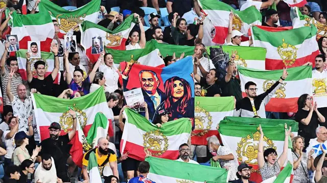 Los aficionados iraníes en las gradas ondean banderas verdes, blancas y rojas, con un escudo dorado en la mitad.