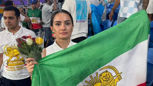 Una mujer joven sostiene un ramo de rosas y una bandera iraní verde, blanca y roja con el sol y el león.