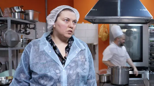 Yulia lleva un mono de trabajo y una redecilla para el pelo en una cocina industrial con paredes naranjas. Un hombre con gorro y delantal de chef camina detrás de ella.