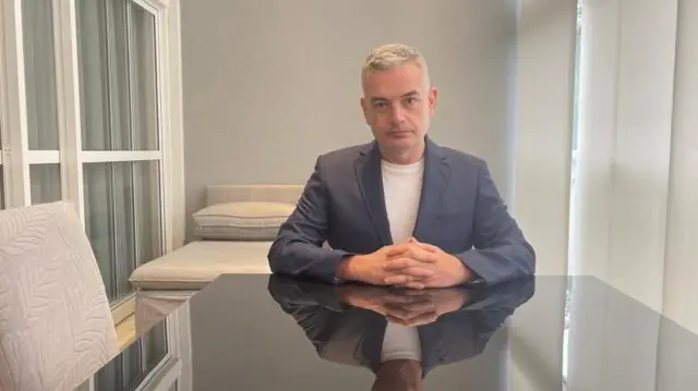 El padre de Victoria está sentado a una mesa de cristal con las manos juntas. Tiene el pelo gris y lleva una chaqueta azul y una camiseta blanca.