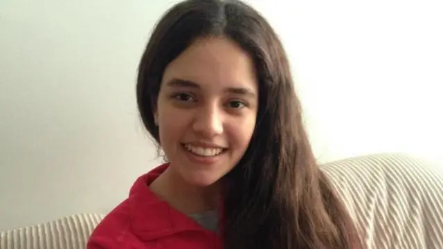 Victoria, vestida con una blusa roja y sentada en un sofá, sonríe a la cámara. Tiene el pelo largo y castaño.