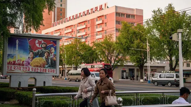 Los peatones pasan junto a un mural (izquierda) que dice "Por la seguridad y el futuro infinito de nuestra nación", mientras que al fondo se ve un edificio adornado con el lema "Viva la gran política que prioriza al pueblo" en Pyongyang el 8 de mayo de 2026.
