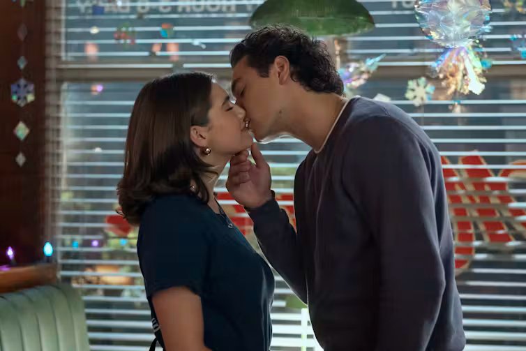 Un chico y una chica se besan en un interior con una ventana cerrada de fondo.