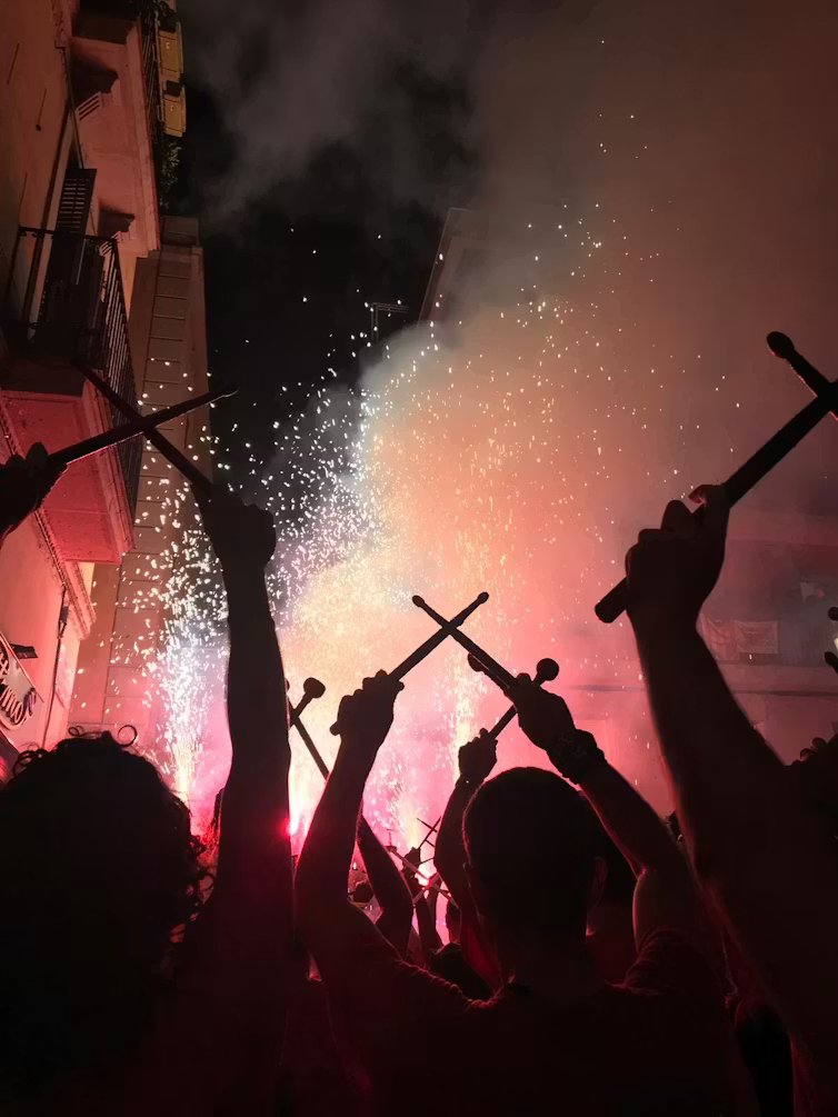Un grupo de tamborileros con las baquetas levantadas por encima de la cabeza, con fuegos artificiales de fondo.