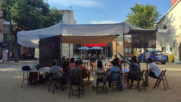 Arquitectura temporal de sombra en una plaza abierta, hecha con palos y telas, bajo la cual tiene lugar una conversación pública con gente sentada en sillas.